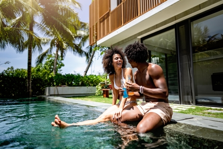 A young couple of African descent enjoying a sunny day at a luxurious poolside, sitting at the pool's edge with drinks in hand. The modern villa in the background enhances the tropical vacation vibe stock photo
