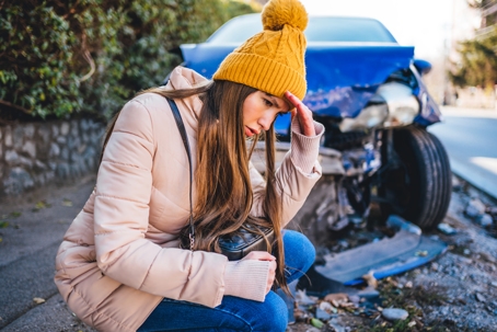 Frustrated woman crouches next to wrecked car after a car accident stock photo
