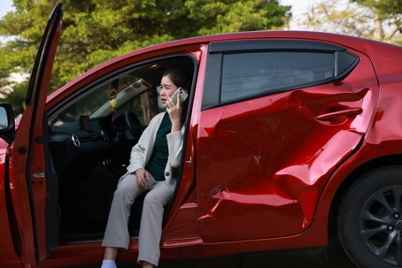 A female motorist is standing on the side of the road after a car accident, assessing the damage to her vehicle