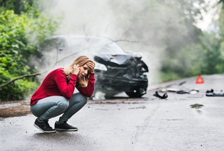 A young woman with smartphone by the damaged car after a car accident, making a phone call.