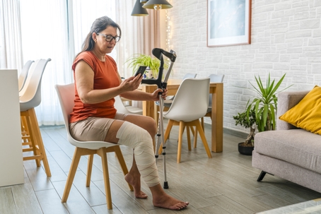 Mature woman with leg in bandage sitting on chair holding crutches using phone for a video call with her doctor or insurance company stock photo
