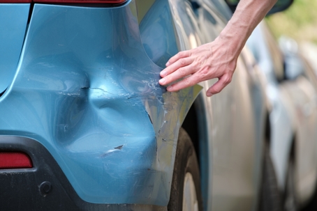 Driver hand examining dented car with damaged fender parked on city street side