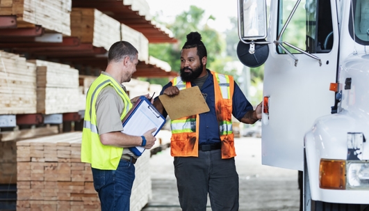 Two multiracial men in lumberyard, with clipboards, truck stock photo
